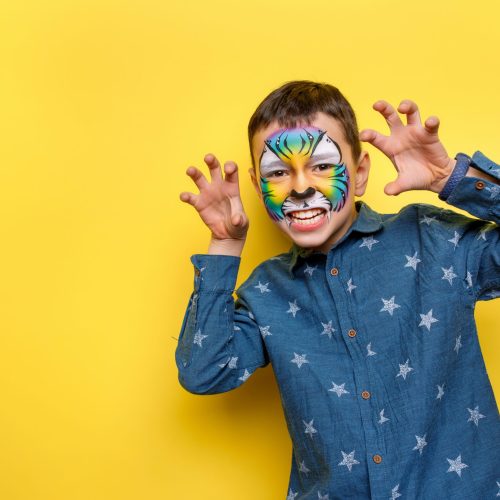 Portrait of ittle cute boy with faceart on birthday party, cute colorful tiger isolated on yellow background.